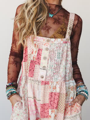 A close-up torso shot of a patchwork overall jumpsuit in soft pinks and creams layered over a sheer dark lace long-sleeve top, styled with stacks of beaded bracelets and a turquoise butterfly pendant — the kind of outfit that looks like it came toget