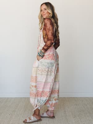 A model glances back over her shoulder in a flowy patchwork-print jumpsuit with a sheer lace long-sleeve underlayer, styled with stacked beaded bracelets and white buckle sandals against a soft studio wall — effortlessly boho and a little unexpected.