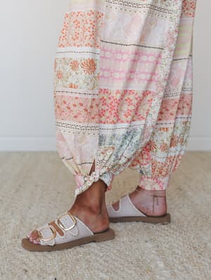 A close-up of flowy patchwork-print pants tied at the ankle, paired with double-buckle slide sandals — all soft pinks, peach, and orange botanicals that feel like a garden in motion.