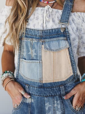 A close-up torso shot of patchwork denim overalls layered over a floral off-shoulder top, styled with stacked beaded bracelets and layered necklaces — the kind of outfit that looks like it was put together effortlessly but definitely wasn't.