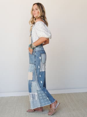 A side-profile shot of a model in patchwork denim overalls styled with a floral off-shoulder puff-sleeve top and studded sandals — the kind of outfit that feels like it was pieced together with intention and a really good eye.