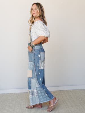 A side-profile shot of a model in patchwork denim overalls styled with a floral off-shoulder puff-sleeve top and studded sandals — the kind of outfit that feels like it was pieced together with intention and a really good eye.