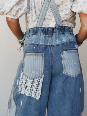 A close-up back view of wide-leg denim overalls layered over a floral blouse, showing off star print fabric panels, striped pocket patches, and a crochet lace appliqué — the kind of jeans that feel like they have a whole personality.