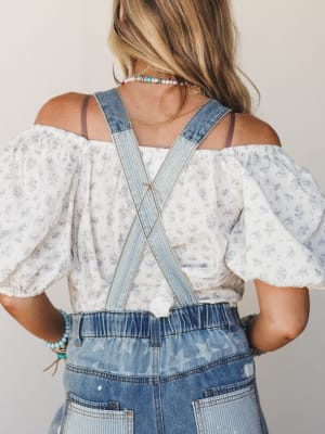 Shot from the back, this look layers a flowy off-shoulder floral top under denim overalls with criss-cross straps and a star print — the kind of outfit that feels effortless and a little nostalgic all at once.