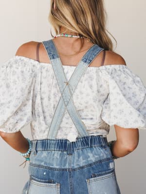 Shot from the back, this look layers a flowy off-shoulder floral top under denim overalls with criss-cross straps and a star print — the kind of outfit that feels effortless and a little nostalgic all at once.
