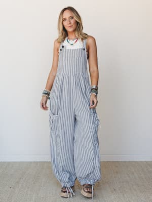 A wide-leg striped overall in soft blue-grey and white pairs with a white tank and stacked colorful bracelets — the kind of effortless, personality-forward outfit you'd wear to the farmers market and somehow look perfectly put together.