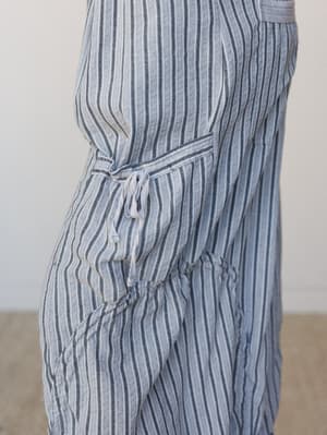 A close-up side view of a striped linen-blend overall, showing the relaxed waist tie and tiered skirt detail — the kind of piece that feels like it was made for slow mornings somewhere coastal.