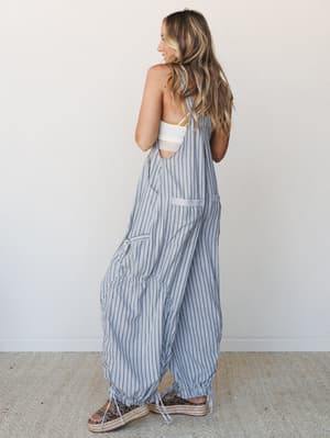 A model shot from behind showing a relaxed, wide-leg striped overall with ruched ankle ties and an open back, styled over a white bralette and paired with espadrille platform sandals — effortlessly laid-back with a summery, free-spirited feel.