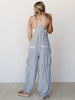 A back-facing model shows off wide-leg striped overalls with ruched ankles and a low-cut back, layered over a mustard lace bralette — casual and a little unexpected in the best way.