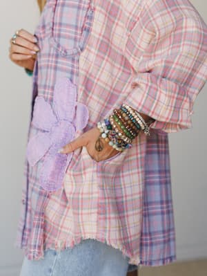 A close-up detail shot of a pastel plaid flannel top with a lavender floral appliqué pocket, frayed hem, and a stacked beaded bracelet arm party that steals the show — equal parts cozy and free-spirited.