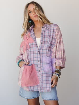 An oversized plaid button-down in pink and lavender gets a handmade-feeling upgrade with appliquéd floral patches, frayed hem, and a contrast pocket — styled over a white tank and denim mini with stacked bracelets for that perfectly undone, expressiv