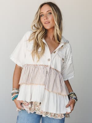 A tiered mixed-fabric top in cream and natural tones layers lace, stripe, and floral patchwork panels with raw fringe edges — styled with stacked beaded bracelets and wide-leg jeans for a free-spirited, lived-in look.