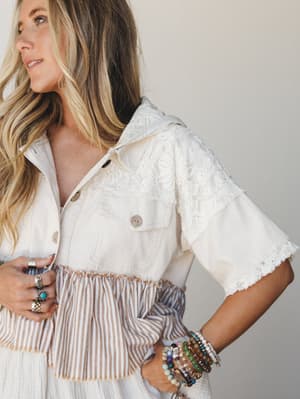 A close-up lifestyle shot of a mixed-fabric cropped jacket in cream and natural tones, featuring lace yoke paneling and a striped peplum hem — styled with stacked beaded bracelets and silver rings for a relaxed, expressive boho feel.