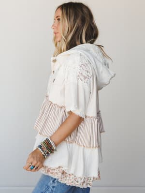 A mixed-fabric hooded top in cream and natural tones, shot in profile — the kind of piece that looks like it collected all its favorite textures along the way: lace, stripe, floral, and gauze, all layered into one very personal outfit.