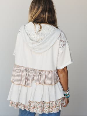 A back-view shot of a cream mixed-media hooded top layered with lace paneling, tiered ruffle hem in stripe and floral prints, styled with stacked beaded bracelets and jeans — effortlessly boho, like something you'd reach for without thinking twice.