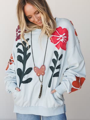 A smiling model wears an oversized sky blue pullover sweatshirt covered in bold floral and leaf appliqué patches in red, burgundy, burnt orange, and dark green — styled with a long chain butterfly pendant necklace and light wash jeans, shot against a