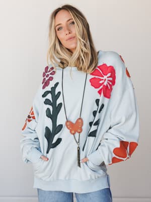 An oversized sky blue pullover covered in bold floral appliqué patches — hibiscus, leafy vines, plum blooms — styled with a long chain butterfly pendant necklace and jeans for an effortlessly expressive, lived-in look.