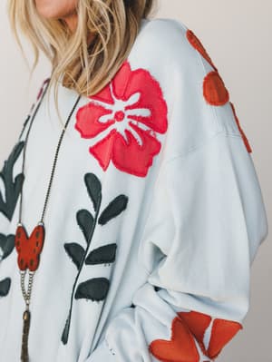 A close-up detail shot of an oversized sky blue pullover covered in bold floral and leaf appliqué patches — red hibiscus blooms, rust orange petals, and dark green foliage — styled with a long layered pendant necklace for an artsy, lived-in feel.