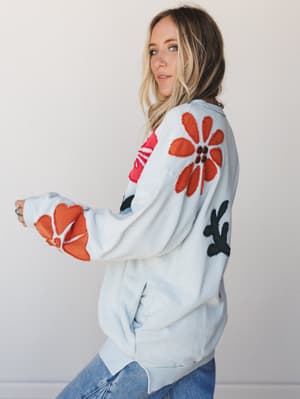 A model shot from the side showing an oversized sky blue pullover sweatshirt covered in bold floral appliqué patches in rust orange, hot pink, and black — the kind of piece that does all the talking while you drink your coffee.
