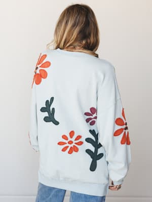 Shot from the back, this oversized sky blue pullover is covered in raw-edge floral and botanical appliqués in rust orange, plum, and deep teal — the kind of sweatshirt that looks like it came from someone's very cool art studio.