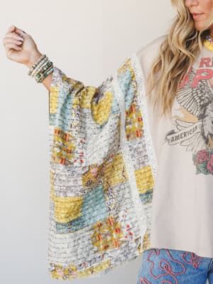 A blonde model holds out a patchwork floral poncho with lace trim, styled over a graphic tee and embroidered denim — the kind of layering piece that looks like it has a whole backstory.