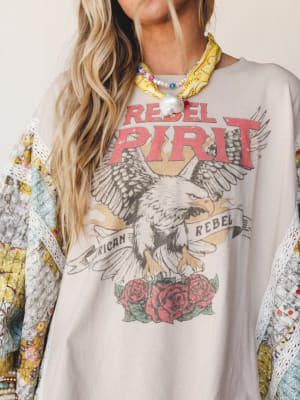 A close-up of a layered boho look centered on an oversized 'Rebel Spirit' eagle graphic sweatshirt in soft tan, styled over a patchwork floral kimono with lace trim and accessorized with a yellow bandana scarf and pearl-and-bead necklace — the kind o
