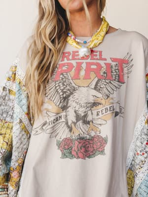 A close-up of a layered boho look centered on an oversized 'Rebel Spirit' eagle graphic sweatshirt in soft tan, styled over a patchwork floral kimono with lace trim and accessorized with a yellow bandana scarf and pearl-and-bead necklace — the kind o