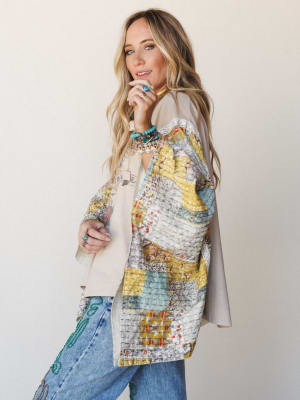 A model styled in a patchwork poncho layered over a graphic tee and embroidered jeans, shot in profile against a soft studio wall — the whole look feels like a well-loved wardrobe that tells a story.