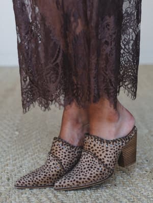 A close-up shot of leopard-spot printed mules with silver stud detailing and a stacked wood heel, styled against the lace hem of a deep mocha dress — the kind of pairing that makes a whole outfit feel intentional.