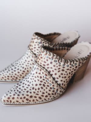 A pair of pointed-toe mules in a cream and brown mini animal print, shot up close on a soft grey surface — the kind of shoe that does the talking so your outfit doesn't have to.