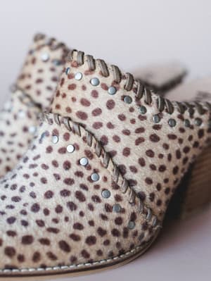 A close-up detail shot of a spotted animal-print mule with silver stud trim, lace-up detailing, and a wood block heel — the kind of shoe that does all the talking.