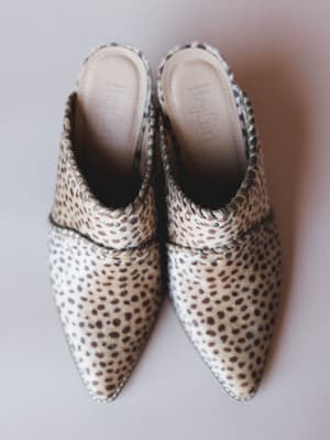 A pair of pointed-toe mules shot from above on a soft lavender-grey surface — the mini cow print texture and structured strap give them that sweet spot between polished and a little wild.