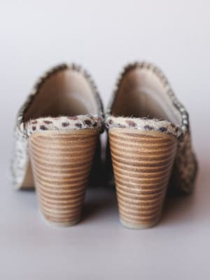 A close-up rear shot of a pair of heeled mules with a fuzzy mini cow-print upper and stacked wood-grain block heels — earthy, a little wild, and exactly the kind of shoe that turns a simple outfit into a personality.