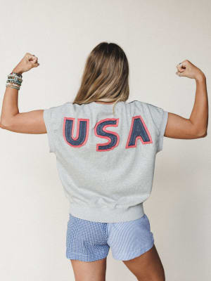 Shot from behind, a model flexes both arms to show off the bold USA appliqué lettering on a heather gray short-sleeve sweatshirt, paired with mixed-pattern gingham and stripe shorts — playful, proud, and ready for a backyard cookout.