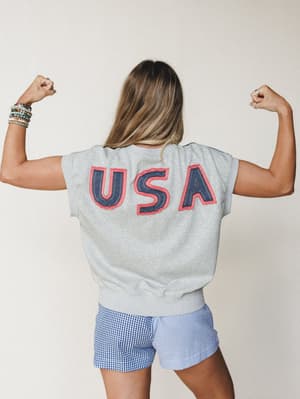 Shot from behind, a model flexes both arms to show off the bold USA appliqué lettering on a heather gray short-sleeve sweatshirt, paired with mixed-pattern gingham and stripe shorts — playful, proud, and ready for a backyard cookout.