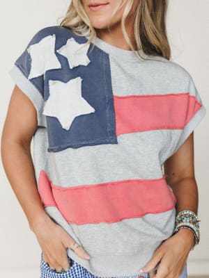 A heather gray short-sleeve sweatshirt top with a faded American flag patchwork design — navy canton with raw-edge star appliqués and coral stripe panels — styled with stacked boho bracelets and a peek of gingham below.