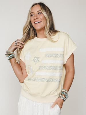 A soft butter yellow sweatshirt-style top with a patchwork flag graphic made from floral fabric strips and chenille star appliqués, styled with white wide-leg pants and stacked beaded bracelets — casual and expressive with a vintage-Americana feel.