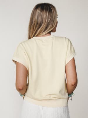 A relaxed, back-facing shot of a cream-toned short-sleeve sweatshirt top styled with a white textured skirt and stacked turquoise bracelets — the kind of effortless warm-weather outfit that looks like it came together in two minutes and somehow looks