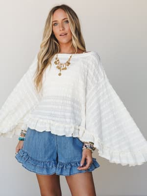 A cream open-knit sweater top with dramatic bell sleeves and ruffle hem is styled with denim ruffle shorts, gold coin jewelry, and stacked bracelets — effortlessly boho and just right for a warm-weather day out.