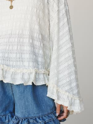 A close-up detail shot of a cream open-knit blouse with ruffle trim at the hem and cuffs, paired with a ruffle-hem denim skirt — soft, romantic, and very much the kind of top you reach for when you want to feel put-together without trying too hard.