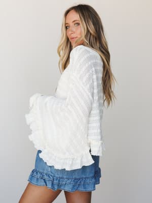 A cream knit top with dramatic ruffle trim and bell sleeves is styled with a denim mini skirt — the kind of outfit that feels effortless but has clearly thought about itself.
