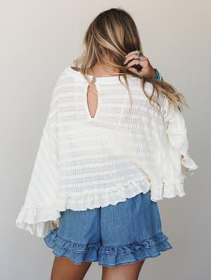 Shot from behind, a model in a cream open-back knit top with dramatic ruffle bell sleeves pairs it with denim ruffle-hem shorts — the whole look has that effortless, slightly undone energy that feels equal parts cozy and romantic.