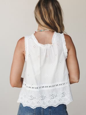 A back-view shot of a white eyelet sleeveless top with a tiered scalloped hem, paired with denim and a colorful beaded necklace — the kind of top that feels like a summer afternoon without trying.