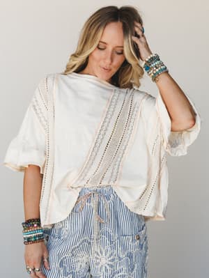 A relaxed, effortlessly boho top in natural cream with lace inset panels and flutter sleeves, styled with blue striped embroidered shorts and stacked beaded bracelets — the kind of outfit that looks like it took zero effort and somehow everything.