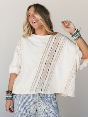 A laughing blonde woman styled in a breezy cream oversized top with diagonal lace inset panels and a peek of blue striped embroidered pants below — the kind of effortless, textured outfit that feels like a good-mood day.