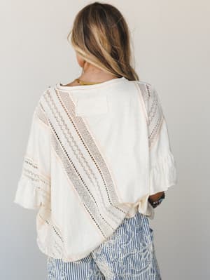 Shot from behind, this cream top with diagonal lace inset panels and ruffle sleeves has an effortless, almost vintage quality — paired with printed blue pants and a beaded bracelet, it feels like a weekend that has nowhere to be.