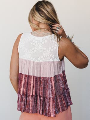 Shot from behind, a tiered sleeveless top layers a white floral lace yoke over a blush waffle-knit middle tier and a richly patterned burgundy boho print ruffle hem — styled with stacked rings and layered gold necklaces for that effortlessly collecte