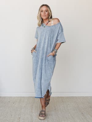 An oversized mineral-washed maxi tee dress in a faded denim blue, styled off-the-shoulder with layered boho jewelry and platform sandals — the kind of effortless look that feels like a good day.