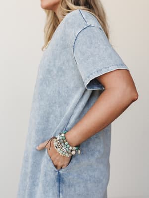 A side-profile close-up of a mineral-washed denim-blue maxi tee dress, hand tucked into a side pocket, styled with a stack of earthy beaded bracelets — effortless and a little collected.
