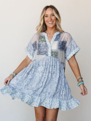 A smiling model twirls a flowy patchwork mini dress in soft blue and mixed prints, styled with stacked turquoise and gold bracelets — the kind of dress that feels like a good mood.